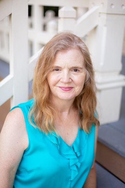 Author Stephanie Weaver in 2021. A white woman with silvering strawberry blonde longer hair, wearing a teal sleeveless top with ruffles at the neckline. She's sitting on the porch of an old home with a white railing behind her.